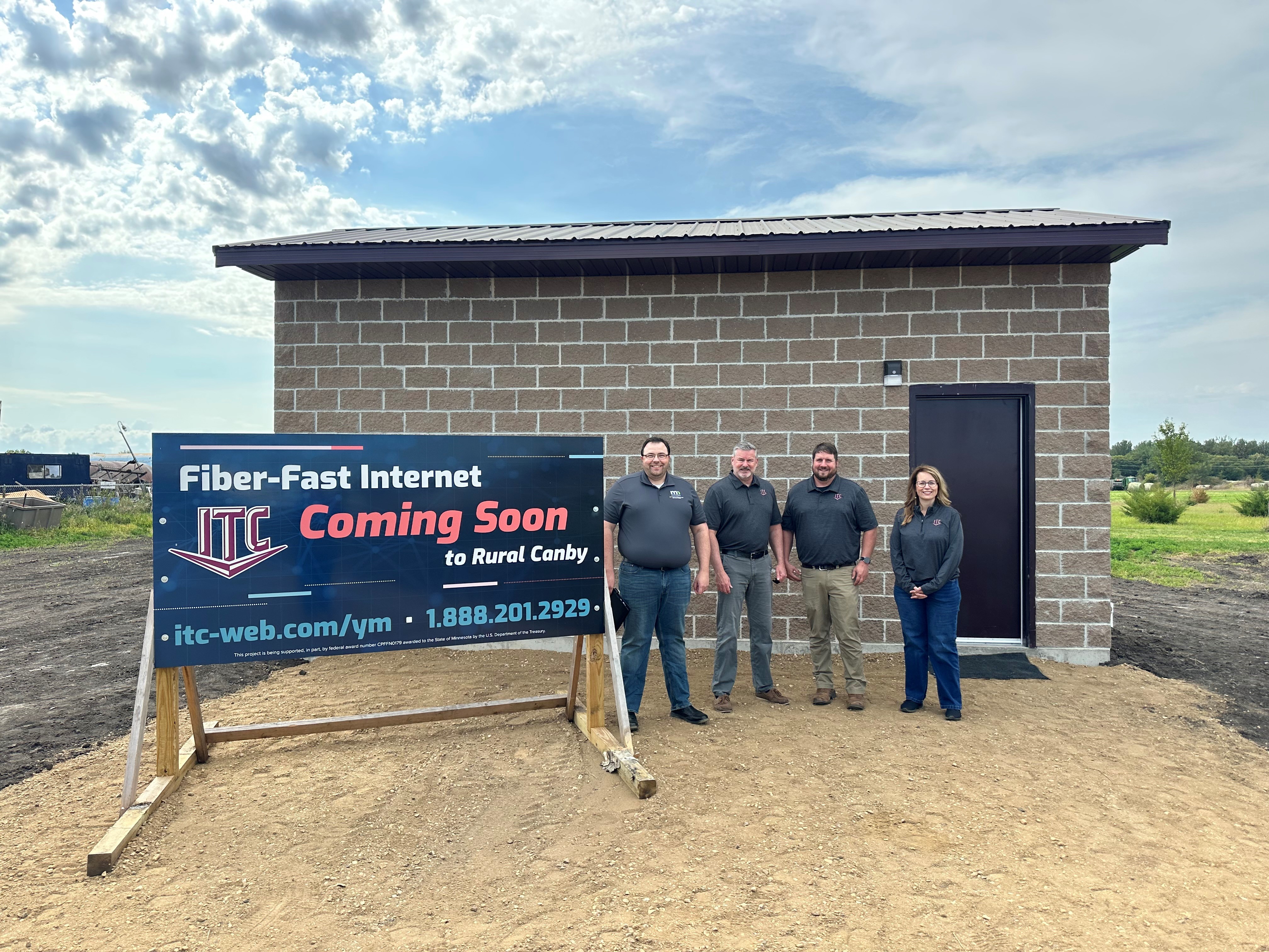 Four people stand in a gravel patch in front of a recently completed brown brick building. There is a sign on the left of the photo that reads 