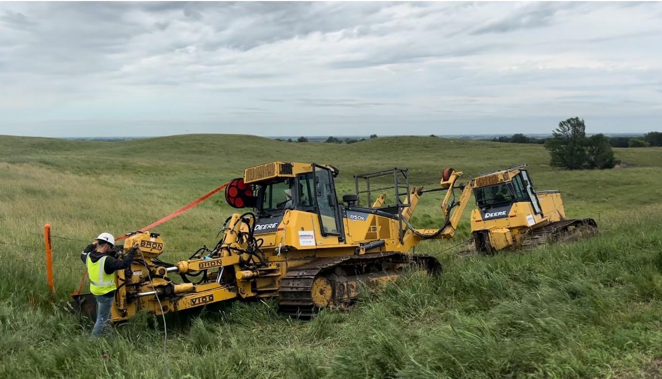 A man stands in a grassy ditch alongside a road with yellow Deere construction equipment.